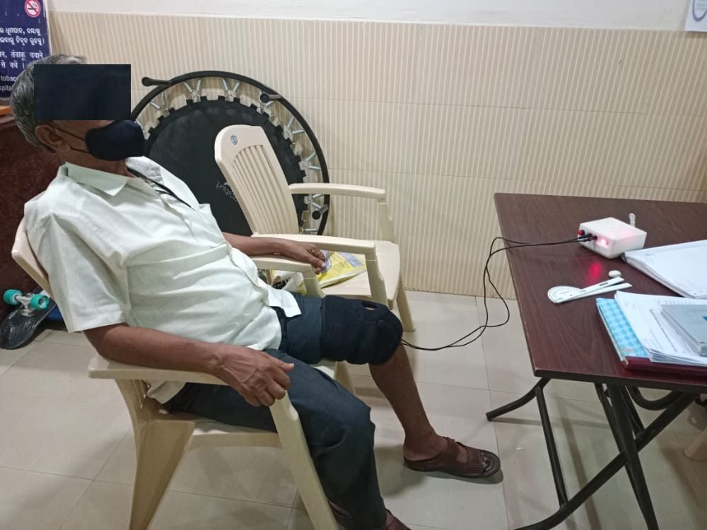 A patient seated in a clinic using the SwaKnee device connected to the control module (identity obscured)
